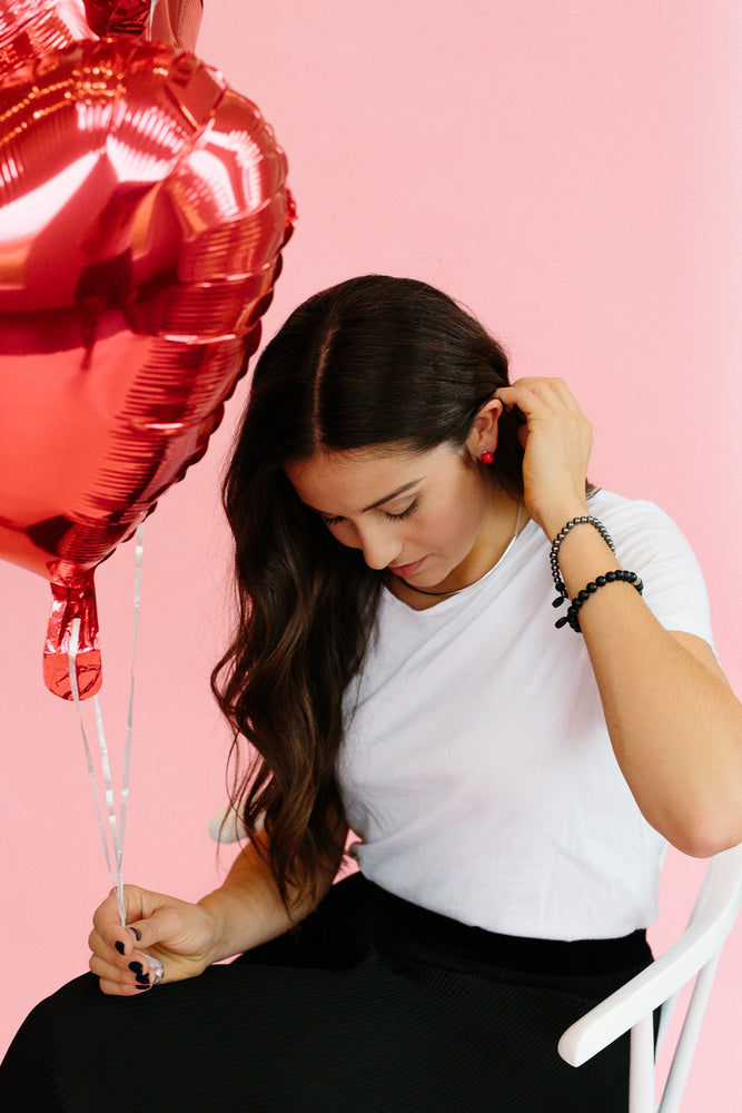 Park and Buzz radiance stud. Sparkle ball earrings. Hillberg and Berk. Canadian Brand. Glitter ball earrings. Red earrings jewelry jewellery. Girl showing earring in white shirt and black pants holding red heart balloons on pink background. Fun photo idea.